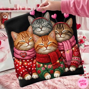May include: Black pillow featuring four smiling cats wearing sweaters and scarves. The cats have headbands with hearts. The sweaters are red and green with heart and rose patterns. One scarf says "LOVE". The pillow is held by a person.