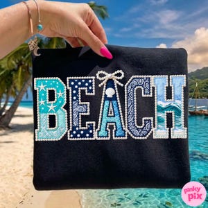 May include: Black sweatshirt with the word "BEACH" embroidered in various shades of blue and white. The letters are decorated with seashells, stars, and patterns, and outlined with white pearls. The background shows a tropical beach scene.