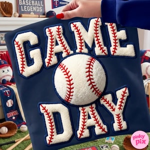 May include: Navy blue pillow with the words "GAME DAY" in white, baseball-style lettering with red stitching. A baseball is centered on the pillow. The pillow is held by a person with red nail polish.