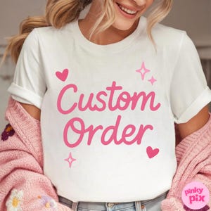 May include: White t-shirt with the words "Custom Order" in pink script, accented with pink hearts and stars. The shirt has rolled-up sleeves. The image also shows a pink cardigan.