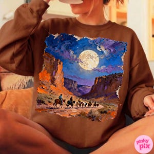 May include: Brown sweatshirt featuring a vibrant, textured painting of a desert canyon scene under a full moon. The artwork depicts cowboys on horseback, with a night sky in shades of blue, purple, and orange. The sweatshirt has a crew neck.
