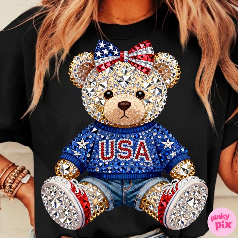 May include: Black t-shirt featuring a rhinestone-studded teddy bear design. The bear wears a blue sweater with "USA" and stars, denim shorts, and sneakers with red and white accents. A bow with an American flag pattern adorns its head.