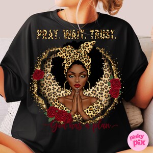 May include: Black t-shirt with a gold glitter background and a black woman with a leopard print headscarf. The text "Pray, Wait, Trust. God has a plan" is printed on the shirt.