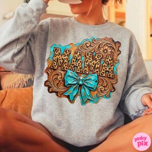 May include: A gray sweatshirt featuring a decorative design with the word "MAMA" in a leopard print font. The design includes a turquoise bow and a brown, textured background.