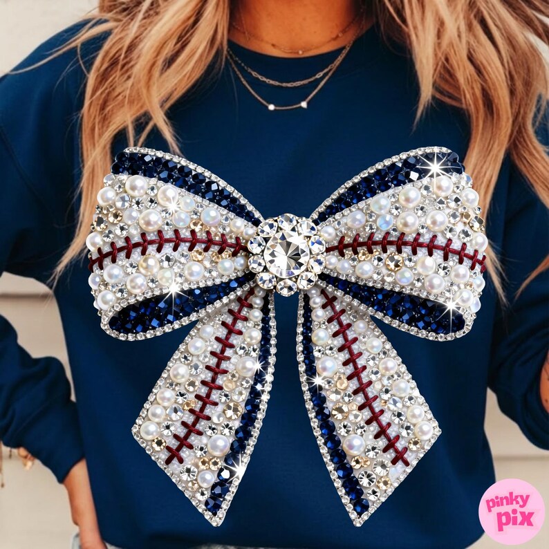 May include: Navy blue sweatshirt featuring a large bow design. The bow is embellished with white pearls, clear rhinestones, and dark blue and red accents, resembling a baseball. A large, sparkling rhinestone is in the center of the bow.