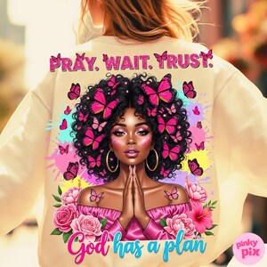 May include: A cream-coloured sweatshirt with a colourful graphic of a woman adorned with butterflies. The text "PRAY. WAIT. TRUST." is at the top, and "God has a plan" at the bottom. Pink and blue splashes of colour enhance the design.