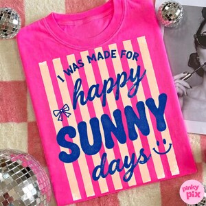 May include: A bright pink t-shirt with the text "I WAS MADE FOR happy SUNNY days" in blue and cream colors. The design includes a bow and a smiling face. The shirt is a crew neck style.