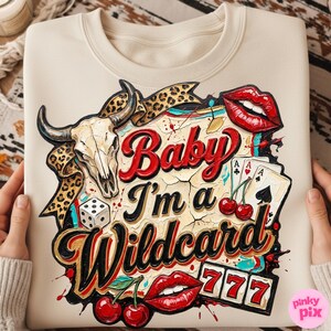 Może przedstawiać: Kremowa bluza z nadrukiem przedstawiającym czaszkę byka, karty do gry, wiśnie i napis "Baby I'm a Wildcard". Projekt zawiera czerwone usta i liczbę 777.