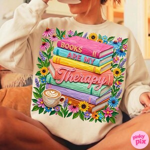 May include: Cream-colored sweatshirt featuring a colorful graphic of stacked books with the words "Books Are My Therapy." The design includes a coffee cup and a floral border with pink, yellow, and blue flowers.