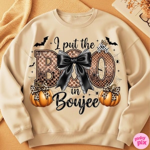 Może przedstawiać: Beżowa bluza z napisem "I put the BOO in Boujee". Projekt zawiera dużą czarną kokardę, pajęczyny, nietoperze i dwie dynie z kokardami w panterkę. Zabawny i świąteczny top z motywem Halloween.