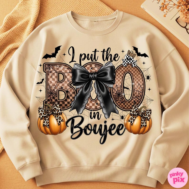 Może przedstawiać: Beżowa bluza z napisem "I put the BOO in Boujee". Projekt zawiera dużą czarną kokardę, pajęczyny, nietoperze i dwie dynie z kokardami w panterkę. Zabawny i świąteczny top z motywem Halloween.