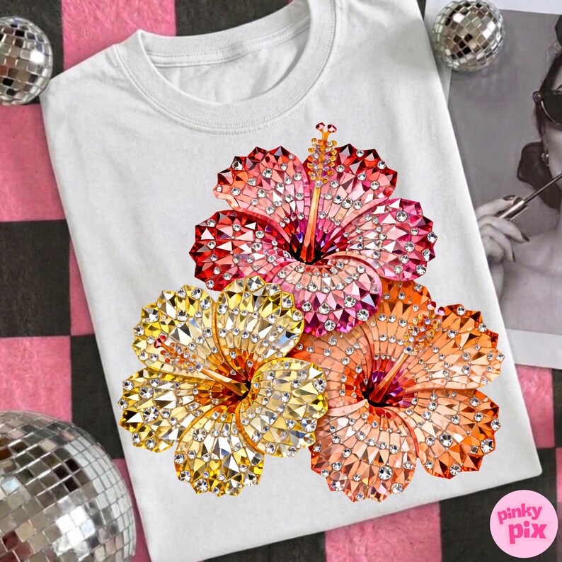 May include: White t-shirt featuring a design of three hibiscus flowers in shades of red, orange, and yellow, embellished with sparkling rhinestones. The flowers are arranged on the front of the shirt. The brand name "pinky pix" is in the bottom right corner.