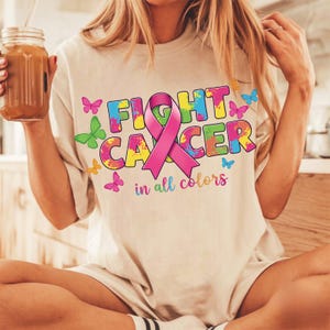 May include: Beige t-shirt with the words "FIGHT CANCER" in colorful block letters and a pink ribbon. The phrase "in all colors" is below the text. Butterflies in various colors are scattered around the text.
