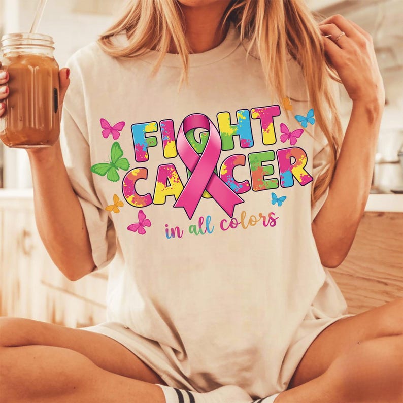 May include: Beige t-shirt with the words "FIGHT CANCER" in colorful block letters and a pink ribbon. The phrase "in all colors" is below the text. Butterflies in various colors are scattered around the text.