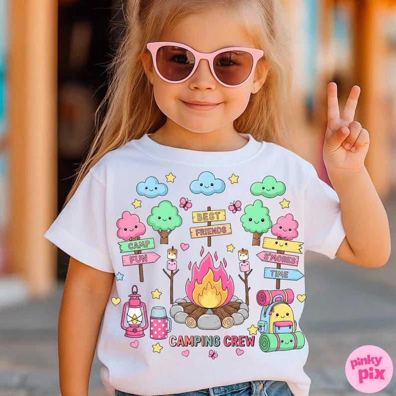 May include: White t-shirt with a colorful camping-themed graphic. The design includes a campfire, trees, clouds, and the words "CAMPING CREW," "CAMP FUN," "S'MORES," and "BEST FRIENDS." The shirt is worn by a child wearing pink sunglasses.