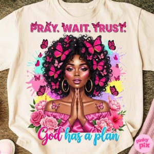 May include: Cream-colored t-shirt featuring a vibrant illustration of a woman with butterflies in her hair and around her. The text "PRAY. WAIT. TRUST." is at the top, and "God has a plan" is at the bottom. Pink and blue accents.
