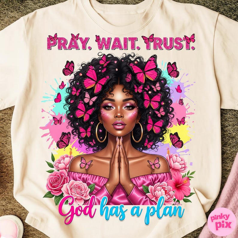 May include: Cream-colored t-shirt featuring a vibrant illustration of a woman with butterflies in her hair and around her. The text "PRAY. WAIT. TRUST." is at the top, and "God has a plan" is at the bottom. Pink and blue accents.