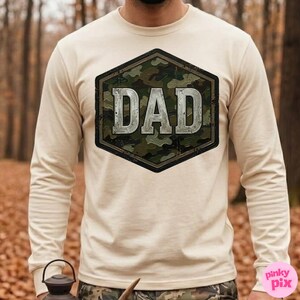 Pode incluir: Uma camisa de manga comprida creme com um design hexagonal de camuflagem. O design apresenta a palavra "DAD" em uma fonte prateada desgastada. A camisa &eacute; feita de um material macio e &eacute; adequada para uso casual.