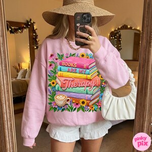 May include: A light pink sweatshirt with a graphic of a stack of books with the words "BOOKS ARE MY Therapy" and a coffee cup. The design is surrounded by colorful flowers.