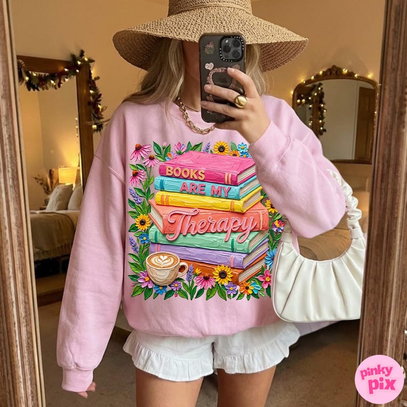 May include: A light pink sweatshirt with a graphic of a stack of books with the words "BOOKS ARE MY Therapy" and a coffee cup. The design is surrounded by colorful flowers.