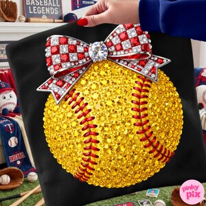May include: A black cushion featuring a softball design covered in yellow rhinestones with red stitching. A bow with red and white checkered accents and a large central rhinestone sits atop the softball. The cushion is held by a person.