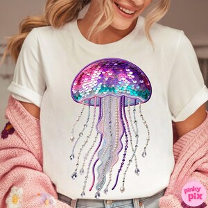 May include: White t-shirt featuring a colorful jellyfish design. The jellyfish has a sequined dome in shades of purple, pink, and teal, with beaded tentacles and dangling crystals. The shirt is a casual, everyday wear item.