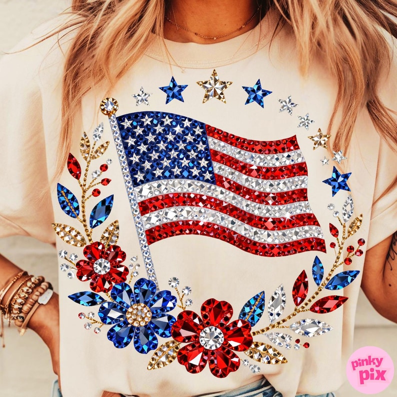 May include: Cream-colored t-shirt featuring a sparkling American flag design. The flag is embellished with red, white, and blue rhinestones. A wreath of flowers and stars surrounds the flag, adding to the patriotic theme. The shirt has short sleeves.