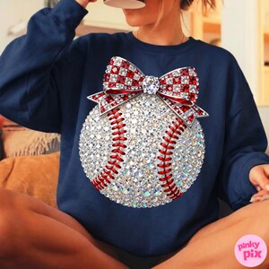 May include: Navy blue sweatshirt featuring a baseball design. The baseball is embellished with sparkling rhinestones and red stitching. A red and white checkered bow with a central jewel adorns the top of the baseball. The sweatshirt is a casual style.
