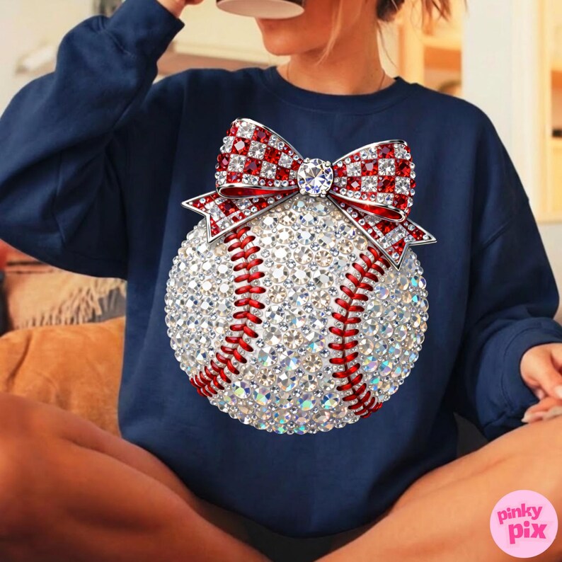 May include: Navy blue sweatshirt featuring a baseball design. The baseball is embellished with sparkling rhinestones and red stitching. A red and white checkered bow with a central jewel adorns the top of the baseball. The sweatshirt is a casual style.