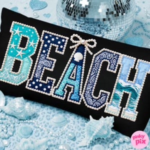 Könnte beinhalten: Schwarzes rechteckiges Kissen mit dem Wort "BEACH" in verschiedenen Blau- und Weißtönen gestickt. Die Buchstaben sind mit Perlen umrandet. Der Buchstabe "B" hat Seestern-Akzente, und der Buchstabe "A" hat eine Muschel und eine Schleife.