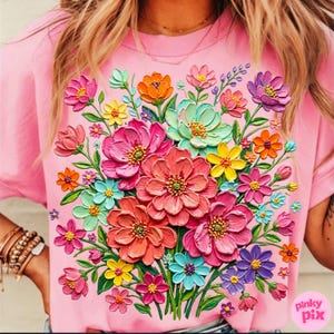 May include: Pink t-shirt featuring a vibrant floral design. The design showcases a bouquet of colorful, textured flowers in shades of pink, orange, yellow, blue, and purple. The flowers are arranged in a dense, layered composition, creating a three-dimensional effect.