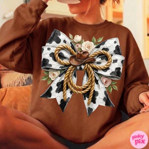 May include: Brown sweatshirt featuring a large bow design with a cow print pattern, a brown cowboy hat, and rope detailing. The bow is adorned with floral accents in white and pink. The sweatshirt is a solid brown color.