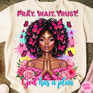 May include: Cream-coloured t-shirt with a colourful illustration of a woman adorned with butterflies. The text "PRAY. WAIT. TRUST." is at the top, and "God has a plan" is at the bottom. Pink and blue accents are used throughout the design.