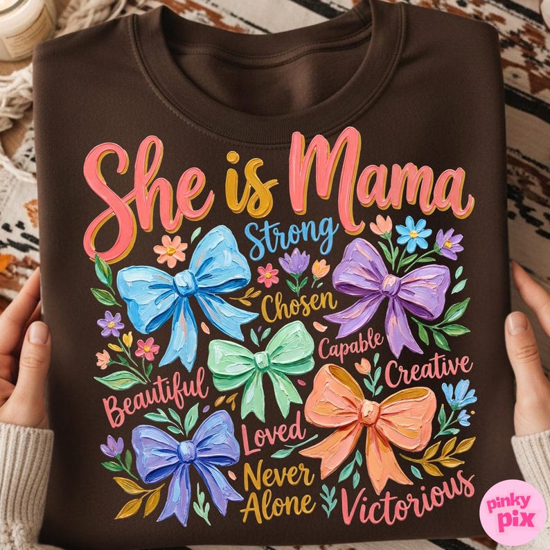K&ouml;nnte beinhalten: Braunes Sweatshirt mit dem Text "She is Mama" in Rosa und Gelb, umgeben von bunten Schleifen und floralen Akzenten. Die W&ouml;rter "Strong", "Chosen", "Capable", "Creative", "Beautiful", "Loved", "Never Alone" und "Victorious" sind ebenfalls enthalten.