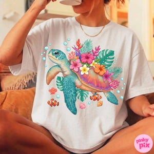 May include: White t-shirt featuring a colorful illustration of a sea turtle adorned with tropical flowers and foliage. The turtle has a blue shell and is surrounded by bubbles and small fish. The shirt has a relaxed fit.