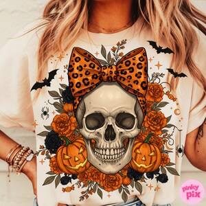 May include: Cream-colored t-shirt featuring a skull with a leopard print bow, surrounded by orange roses, black flowers, and jack-o'-lanterns. Bats, spiders, and candy corn accents complete the Halloween design.