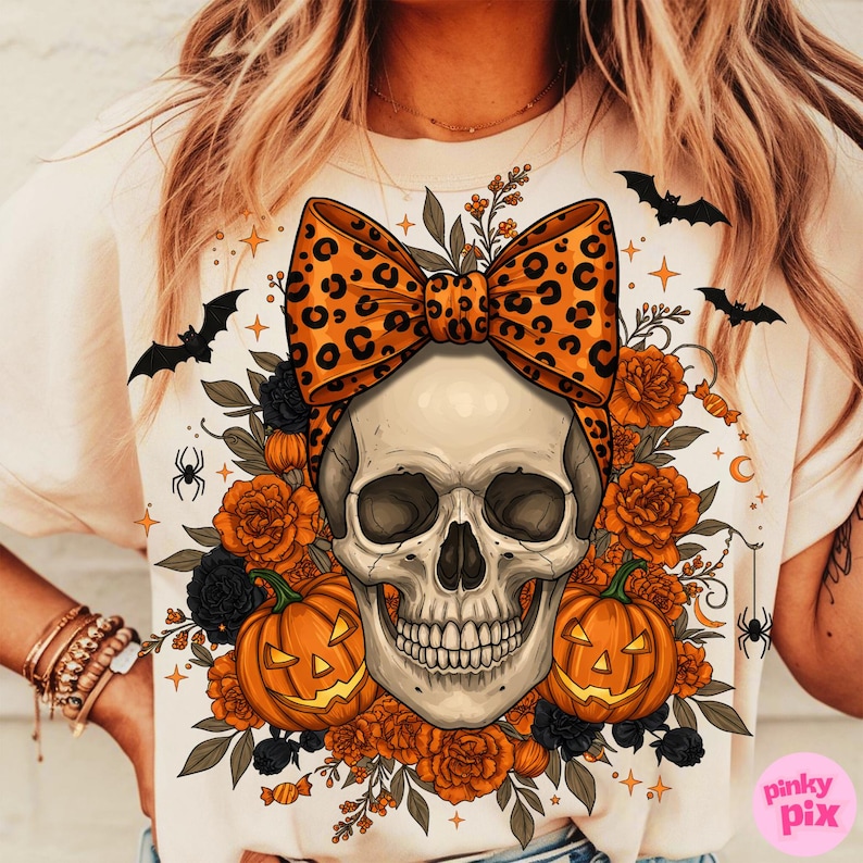 May include: Cream-colored t-shirt featuring a skull with a leopard print bow, surrounded by orange roses, black flowers, and jack-o'-lanterns. Bats, spiders, and candy corn accents complete the Halloween design.
