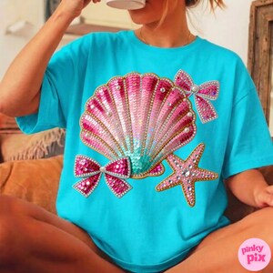 May include: A turquoise t-shirt featuring a sequined seashell design with pink, gold, and teal accents. The design includes a starfish and two bows, all embellished with rhinestones. The shirt is a casual, oversized fit.
