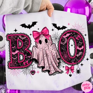 Puede incluir: Una impresi&oacute;n blanca con tem&aacute;tica de Halloween con la palabra "BOO" en rosa, con un fantasma en el centro. Las letras est&aacute;n decoradas con murci&eacute;lagos, telara&ntilde;as y encaje. Lazos rosas y una ara&ntilde;a completan el dise&ntilde;o.