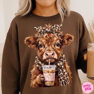 May include: Brown sweatshirt featuring a cow illustration. The cow wears a leopard print scarf and headband, and sips from a coffee cup with a straw. The illustration has a pink cup holder. The sweatshirt is a solid brown color.