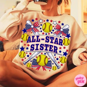 May include: Beige sweatshirt with a softball-themed graphic. The design includes yellow softballs, baseball bats, red, white, and blue bows, and stars. The text "ALL-STAR SISTER" is in blue. The sweatshirt is a casual, sporty style.