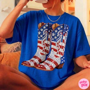 May include: A royal blue t-shirt featuring a pair of cowboy boots. The boots are decorated with red and white stripes, blue stars, and silver embellishments. The boots have brown heels and are a patriotic design.
