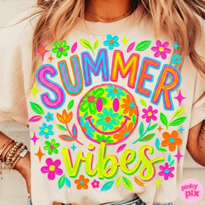 May include: A light beige t-shirt with a colorful graphic design. The design features the word "SUMMER" in pink, blue, and orange, and the word "VIBES" in neon green. A smiley face with flower details is in the center.