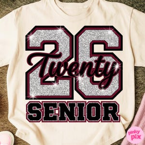 May include: Cream-colored t-shirt with a glittery silver "26" design outlined in black and maroon. The word "Twenty" is in maroon script, and "SENIOR" is in a black, block font. A pink item is visible in the lower right corner.