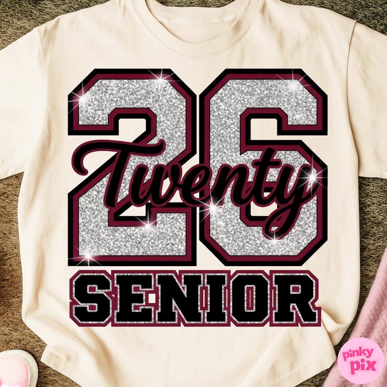 May include: Cream-colored t-shirt with a glittery silver "26" design outlined in black and maroon. The word "Twenty" is in maroon script, and "SENIOR" is in a black, block font. A pink item is visible in the lower right corner.