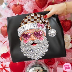 May include: A black pouch featuring a decorative mosaic-style Santa Claus graphic. The Santa design is embellished with sparkling crystals, including pink heart-shaped sunglasses, a checkered hat with a small pink bow, and a white crystal beard.