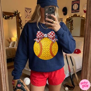 May include: A navy blue sweatshirt with a sparkling yellow softball design, red stitching, and a red and white bow. The image includes red shorts, a baseball glove, and a baseball bat, indicating a baseball theme.