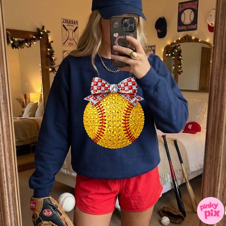 May include: A navy blue sweatshirt with a sparkling yellow softball design, red stitching, and a red and white bow. The image includes red shorts, a baseball glove, and a baseball bat, indicating a baseball theme.