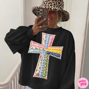 Op de afbeelding: Zwart sweatshirt met een kleurrijke kruisapplicatie. Het kruis is gemaakt van verschillende stoffen met patronen, waaronder bloemen, ruitjes en stippen, en is omrand met kleine witte parels. De sweatshirt heeft lange mouwen.