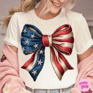 Puede incluir: Camiseta blanca con un gran lazo rojo, blanco y azul. El lazo est&aacute; dise&ntilde;ado para parecerse a la bandera estadounidense.